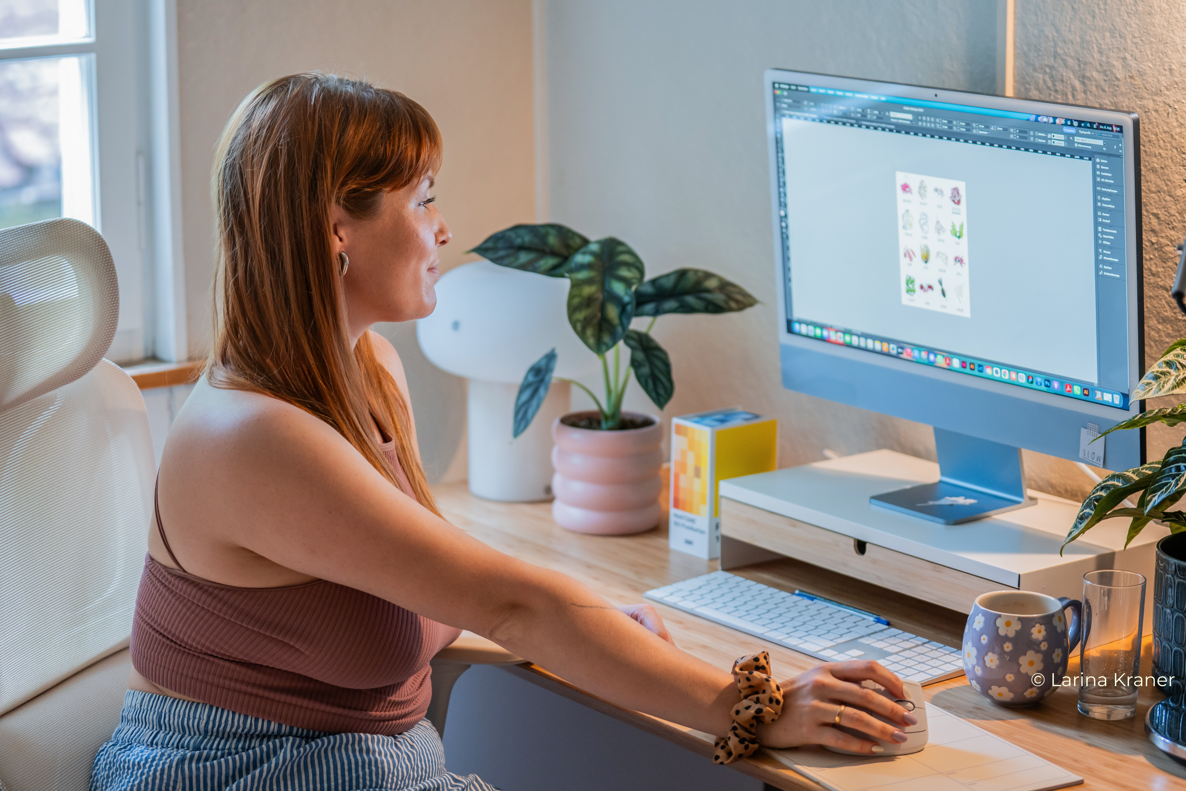 Designerin der Website sitzt an einem Schreibtisch und bearbeitet eine Datei, im Hintergrund Büroeinrichtung und Zimmerpflanze