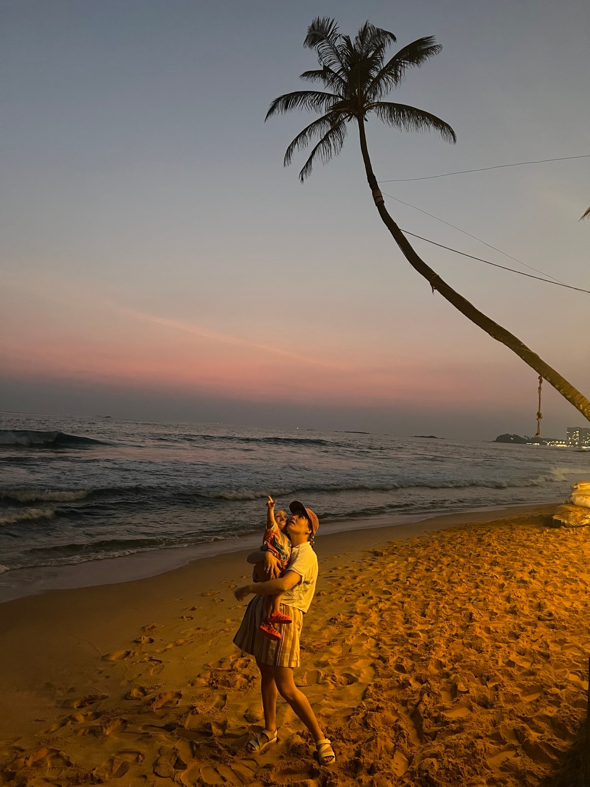 Bild der Designerin mit ihrem Kind auf dem Arm, das in den Himmel zeigt am Strand, vor einer Palme, Abendstimmung