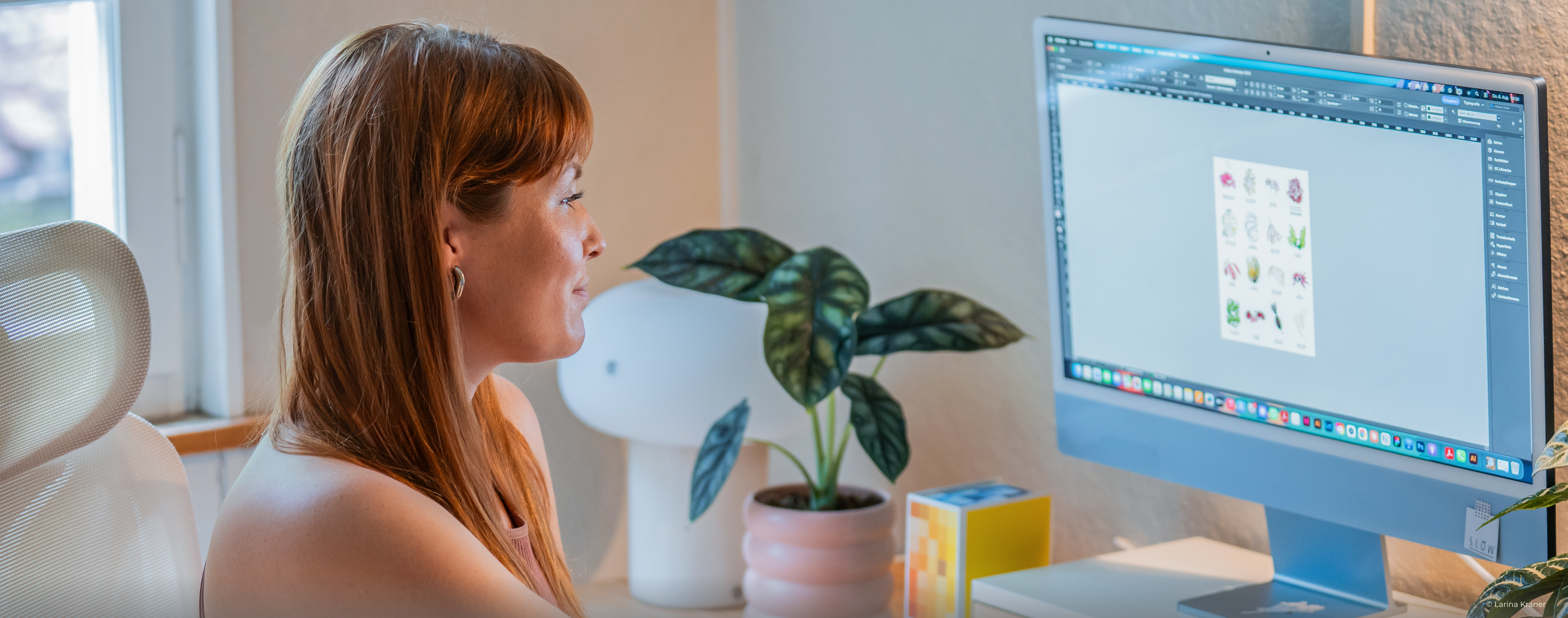 Designerin der Website sitzt an einem Schreibtisch und bearbeitet eine Datei, im Hintergrund Büroeinrichtung und Zimmerpflanze